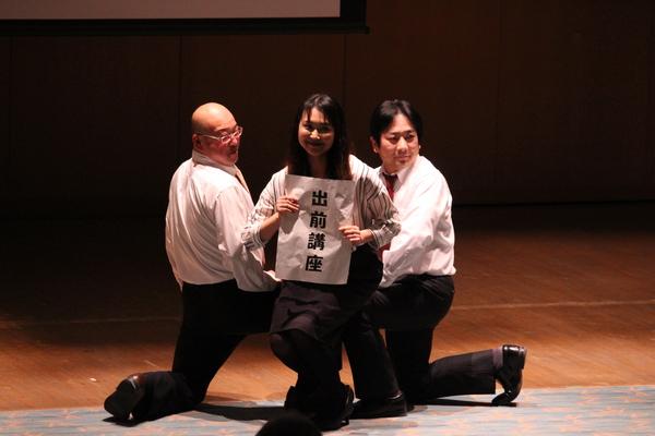 舞台にて「出前講座」と書かれた紙を持って座ろうとしている女性を両脇の男性職員2名が片膝をついて支えている写真