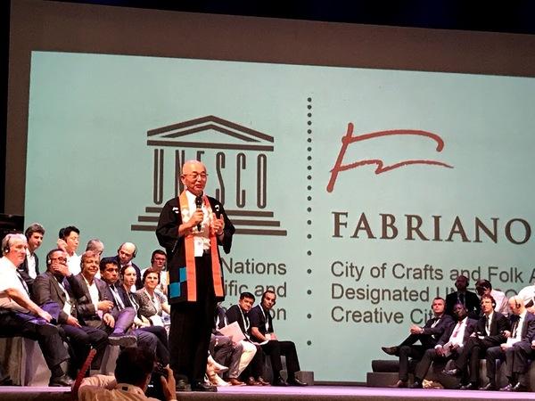 法被を着た市長が舞台で発表していて舞台内の後方で聞いている人々を写した写真