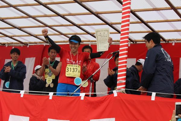 西村広和さんが首に手作りのメダルをかけ、手には賞状を持ちガッポーズして喜んでいる写真