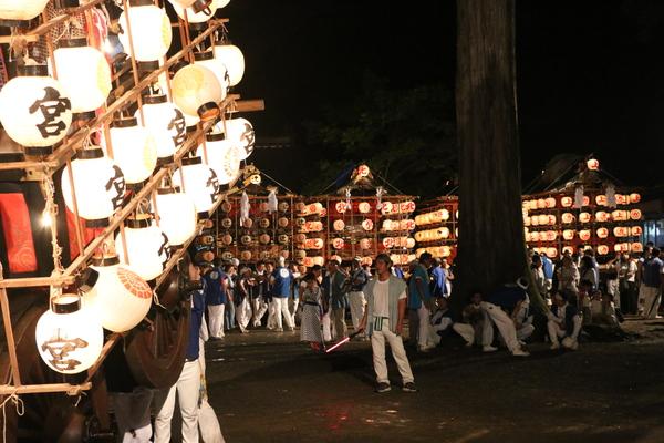 神社の境内に提灯が灯った神輿が数台置いてあり法被姿の男性らが集まっている手前で提灯が灯った山車が上げられている写真