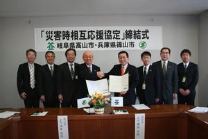 「災害時相互協定」締結式の横断幕の前で、協定書を持ってオレンジ色のネクタイをしている男性と市長が握手をしている記念写真