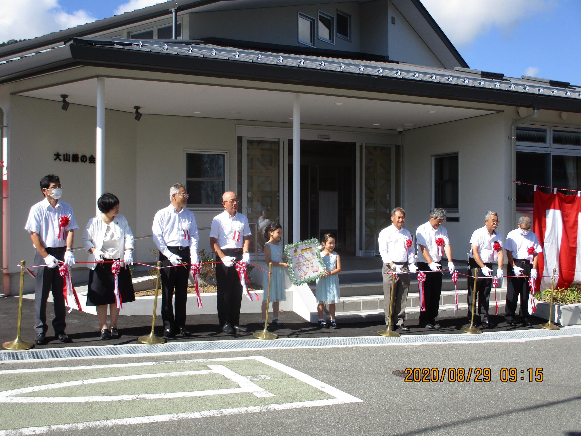 施設前でテープカットを行う式典出席者、中央には「祝」というプレートを持ったこどもが2人
