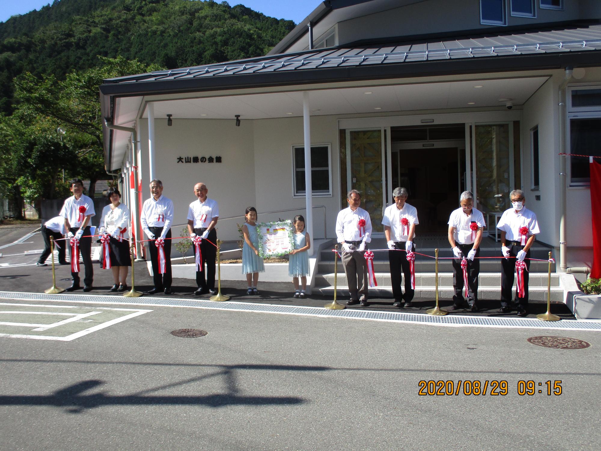 施設前でテープカットを行う出席者、中央に「祝」というプレートを持ったこども2人