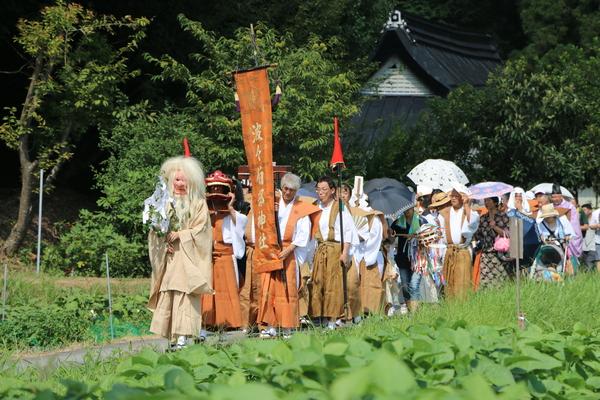 猿田彦のお面を被り玉串を持った人を先頭に神社ののぼり旗を持った袴姿の男性や日傘をさした女性らが道を練り歩いている写真