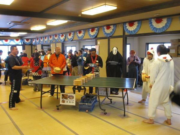 卓球台の右側に白い鳥の衣装を着た二人組がおり、相手チームにボールを打とうボールを手のひらに乗せており、中央には「かおなし」の衣装を着た人が立っている写真