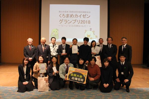 舞台にて、くろまめカイゼングランプリのチームが賞状と全国大会出場切符を手に持ち市長らと一緒に記念撮影している写真