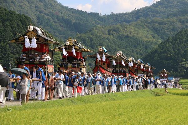 子供らが乗っている山車が数台連なり法被姿の男性らが押して練り歩いている写真