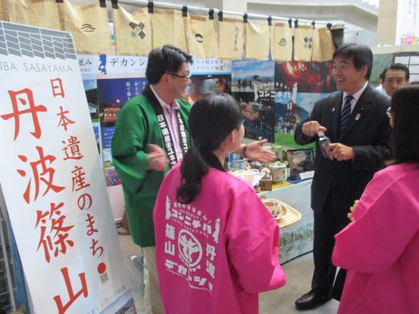 緑と、ピンクの法被を着た男女が、かごやトレーの上の置かれた試食品を持ち、馳文部科学大臣と話をしている写真