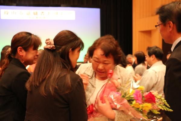 現職員の間を通り、花束を持った退職者の女性が涙しながら職員の方と握手をしている写真