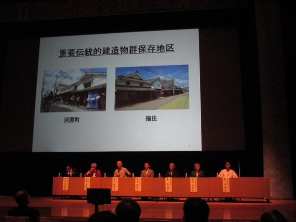 プロジェクトスクリーンに映る2枚の写真と、その前に着席する関係者の写真