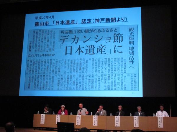 プロジェクトスクリーンに大きく映し出されたデカンショ節の新聞記事と、その前に着席する関係者の中でマイクを持って話す市長の写真