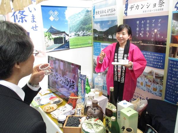 ピンクの法被を着た女性が、小さい紙コップをのせたトレーを持ち、スーツの男性と話をしている写真