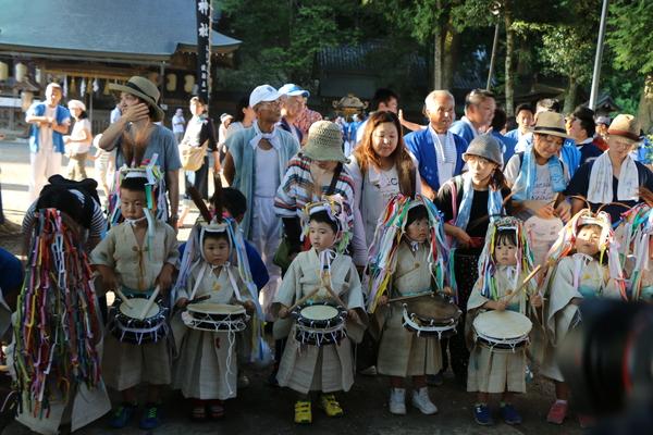 神社にて子供らが飾りのついた帽子を被りベージュの衣装姿でお腹の位置で太鼓を叩いている様子を見守る母親たちの写真