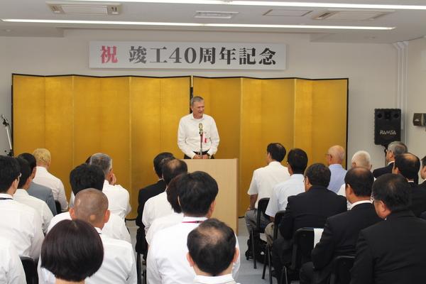 「祝 竣工40周年記念」と書かれた横断幕と金屏風の前で、ノバルティスのダーク・コッシャ社長さんが話をしており、出席者が椅子に座って話を聞いているのを後方から写している写真