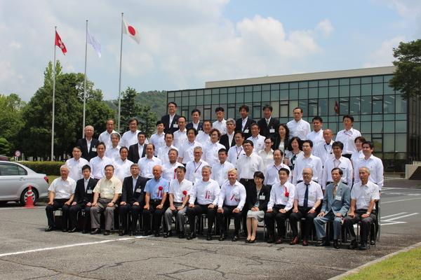 ガラス張りの建物の前で、式典の出席者が集まって並んで写っている記念写真