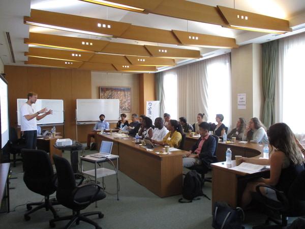 プロジェクトスクリーンの前に立ち、話をする外国人男性をみる、各国の参加者の写真