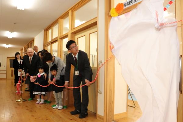 教室の前の名前のプレートに白い布がかけられ、布に紅白の紐がついており、入所した2人の子どもとご家族が紐を引いて除幕式を行っている様子の写真