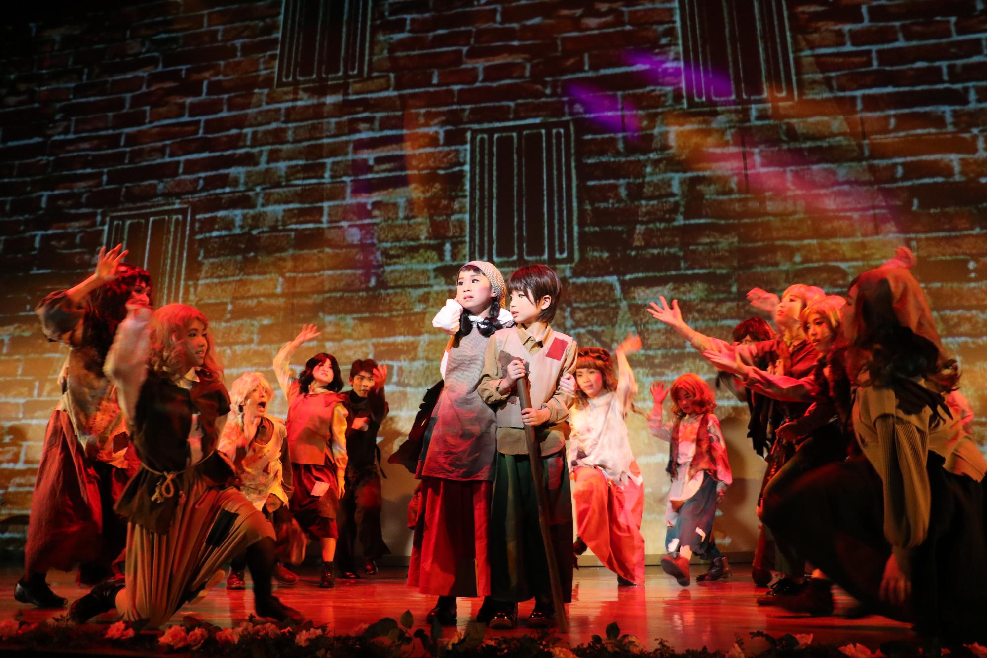 レンガの背景の前で幼いころのサリバン先生と弟が不安げな顔をしている周りに複数の子どもたちがいる写真