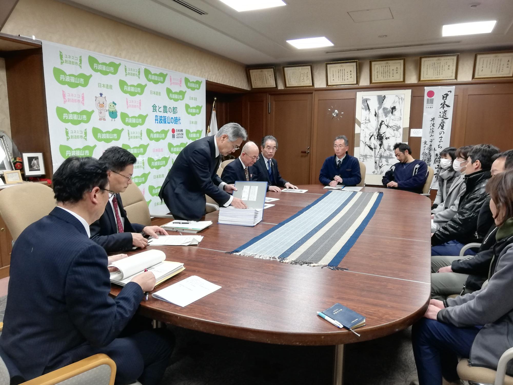 応接室で、市の職員と市民の方々が座り、教育長が立って署名が綴ってあるファイルを見ている写真