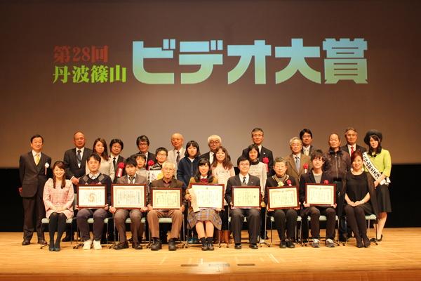 ビデオ大賞で表彰されたグループが舞台の上で額に入った賞状を持って市長らと一緒に記念撮影している写真