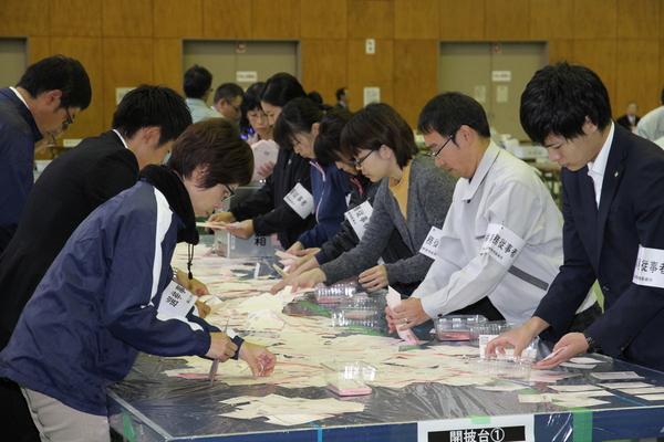 腕に選挙の腕章を付けた係の人が机に広げられた開票用紙を整理している様子の写真