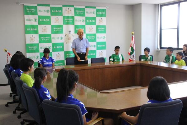 市長が立って話していて、聞いているユニホームを着た、篠山ジュニアホッケークラブの子供達の写真