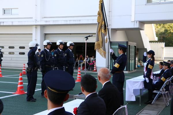 ヘルメットを被った4名の警察官の前に椅子から立ち敬礼している熊谷 奈美さんとその様子を見ている関係者の方々の写真