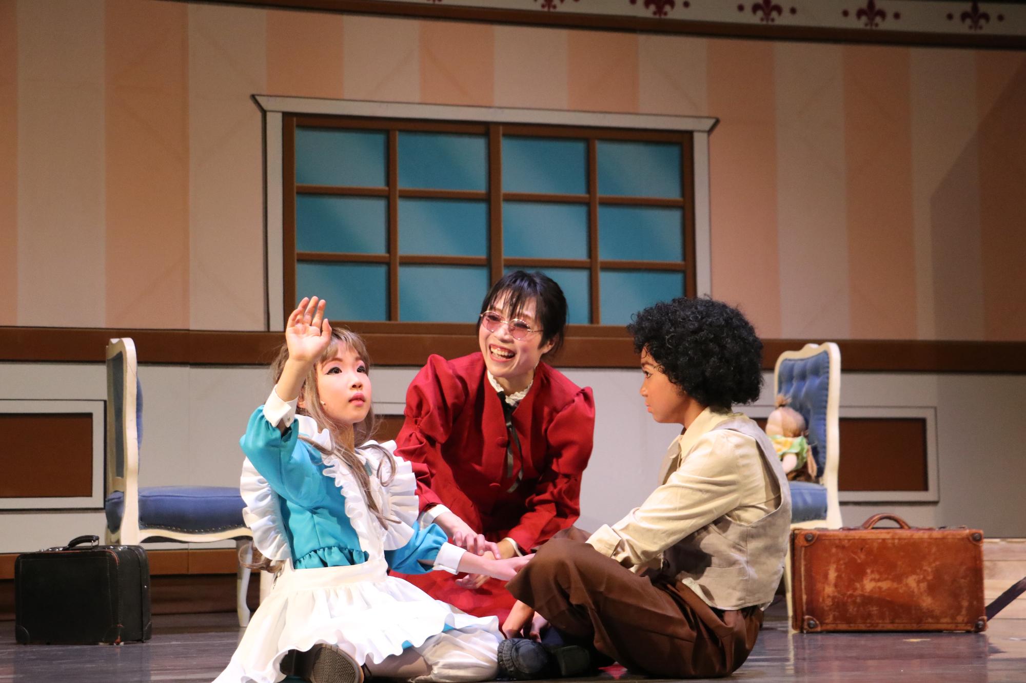 アフロのような髪の子どもが地面にあぐらをかいて座っていて、その正面にヘレンが右手をあげて指文字の練習をしており、笑顔でヘレンの隣でその様子を見つめるサリバン先生の写真