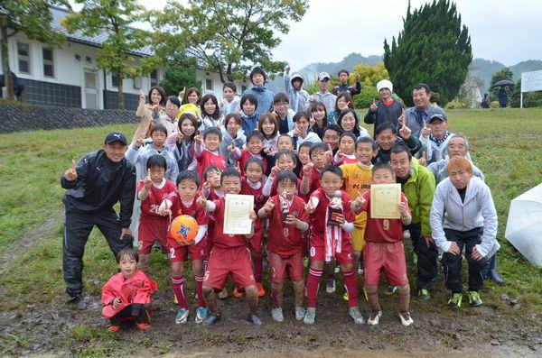 20人ほどのサッカーのユニフォームを着た少年たちとその後ろに保護者の人達が並んでいて、中央の少年がトロフィーと賞状を持っている写真