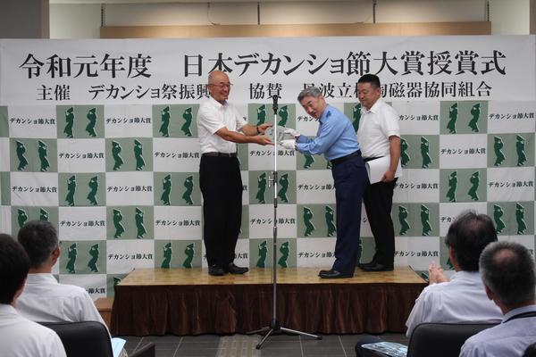 市長から、笑顔で丸いプレートを受け取る森江警察署長、その与野に立つ山内さんの写真