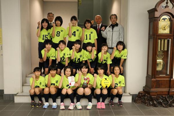 市役所の階段で「Colorsささやま」16名のメンバーと右後方にコーチと市長が全員で右手を曲げて拳を握るポーズを取り記念撮影をしている写真