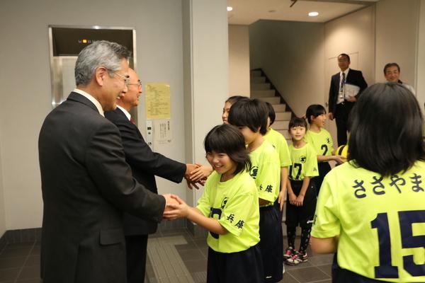 小学生女子のドッチボールチーム「Colorsささやま」のメンバーがユニホーム姿で市役所を訪問し照れながら市長と握手を交わしている写真