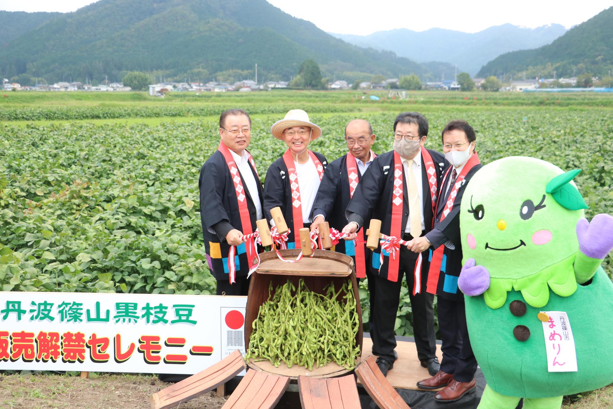 畑の前で、来賓5名が黒枝豆の入った樽の鏡開きしている。そばにはまめりんがいる。