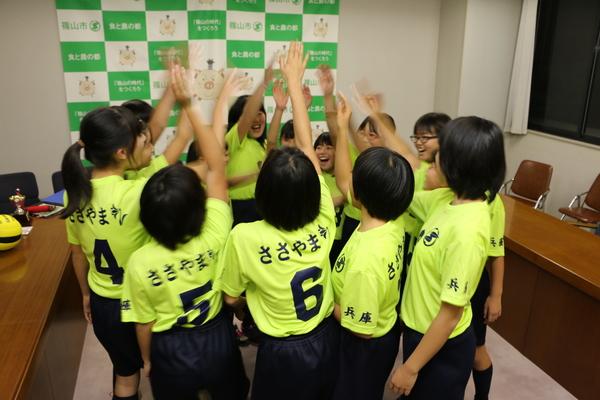 「Colorsささやま」のメンバー全員が市長室で片手を上げて笑顔で気合を入れている写真