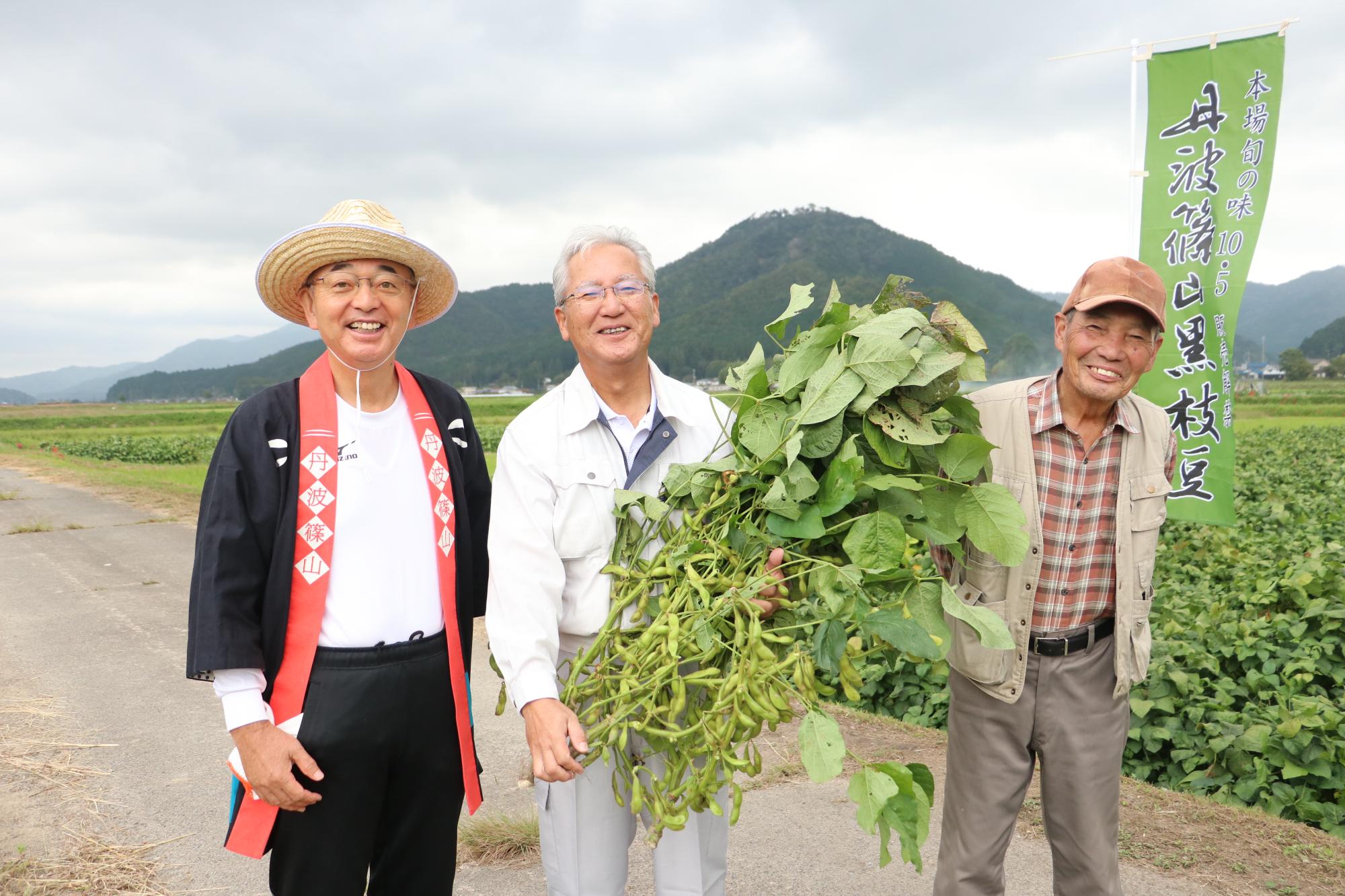 圃場の持ち主の梶村さんが枝豆を手に、お父様と市長と並んで写っている