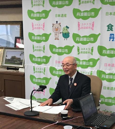 市長がオンラインでシンポジウムに参加している。机には資料が置かれている。