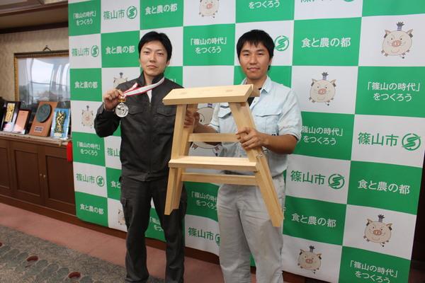 園田 雄一さんが銀メダルを首にかけ、小林 俊文さんが四方転び踏み台を持っている写真