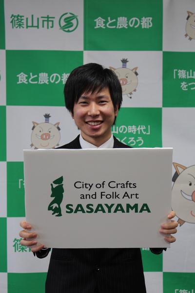 職員の羽馬 雅人さんがユネスコロゴマークを持ち、笑顔で撮影している写真
