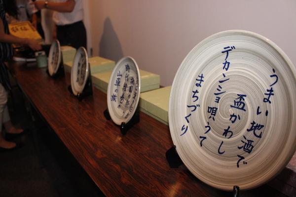 机の上に並べられた、デカンショ節「うまい地酒で盃かわしデカンショ唄うてまちづくり」が書かれたプレートの写真