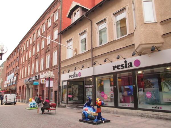 建物がたくさん並び、resiaと看板にかかれた店舗の前に青の恐竜のような置物がありその横にある赤いベンチで女性2人がお話ししている写真