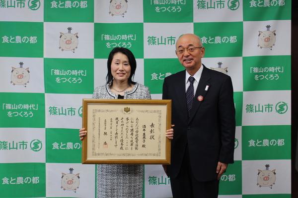 酒井 美奈子先生が額に入っている表彰状を胸の前に持ち、市長と一緒に写っている写真