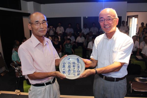 市長と波部さんが、波部さんのデカンショ節「夏は波々伯部祇園の祭り時代絵巻の山車の旅」が書かれたプレートを一緒に持っている写真