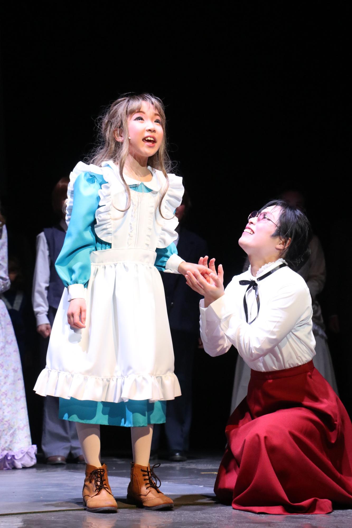 水色の膝下丈のワンピースの上に白いエプロンをつけたヘレンが立って斜め上を見つめ、片膝を地面につけて膝立ちをしているサリバン先生がヘレンの左手を両手で包んでいる写真