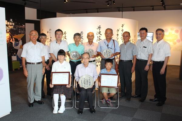 受賞者が、それぞれプレートや、額に入った賞状を持ち、周りに関係者が並ぶ集合写真