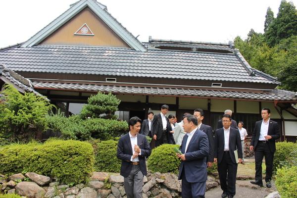 梶山 弘志大臣が古い建物の前の庭園で説明を受けていてその後ろから歩いてくる人々の写真