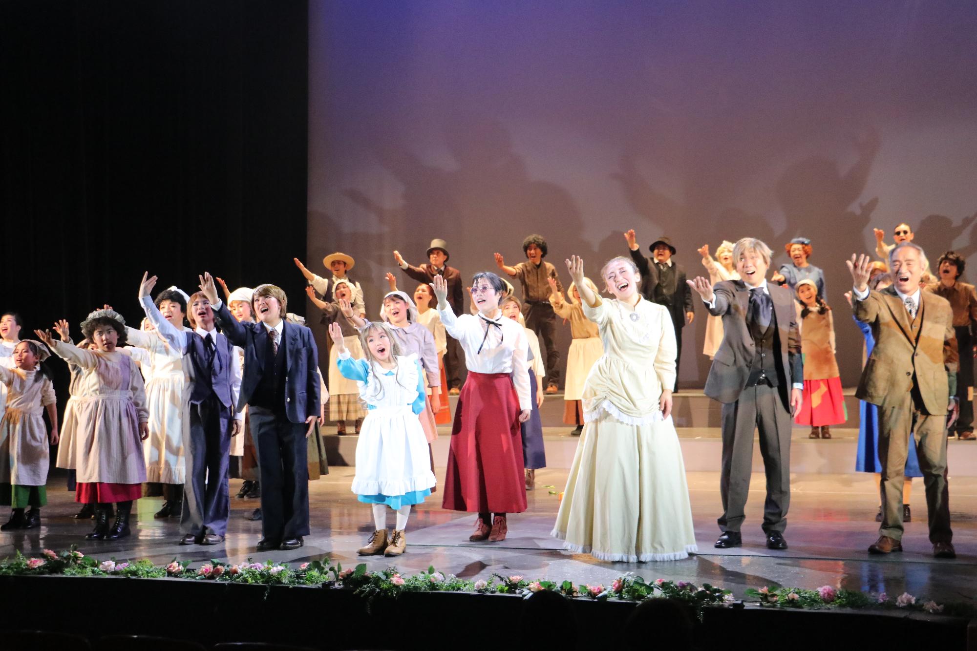 真ん中にヘレンとサリバン先生がおり、その周りにドレス姿の女性や背広姿の男性など出演者が等間隔にならび、右手を斜め上にあげ歌を歌っている写真