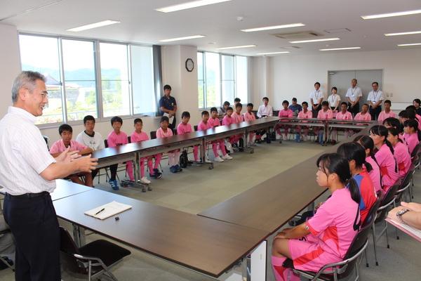 ピンク色のユニフォームを着た篠山丹南ホッケー部（男、女）が会議室に座っており、教育長の話を真剣に聞いている様子の写真