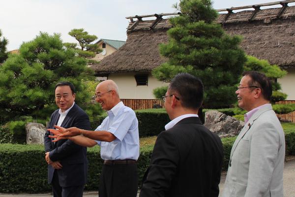 茅葺屋根の建物がある庭の前で梶山 弘志大臣と市長らが談笑している写真