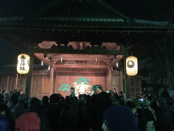 春日神社の能舞台に灯りの灯った提灯が下げられており元朝能を観客が見ている写真