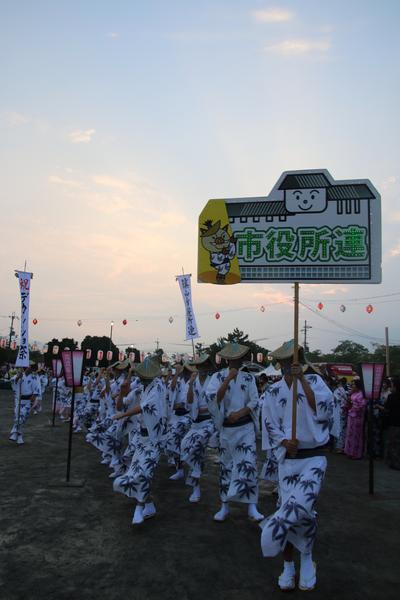 市役所連のプラカードをもった人を先頭に浴衣姿の男性職員の人達が踊っているのを前からうつした写真